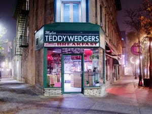 Street view of Teddywedgers storefront with bright lights, glass door, and breakfast sign on a quiet sidewalk.