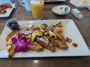 Golden pancakes topped with bananas, nuts, and a purple flower, served with syrup and orange juice.