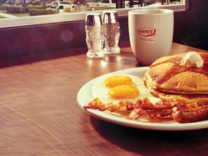 Plate of pancakes, eggs, bacon, and sausage with a Denny's mug on a sunny diner table.