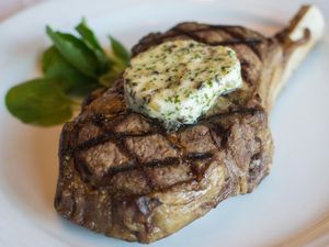 Grilled bone-in steak topped with herb butter, served on a white plate with fresh greens.