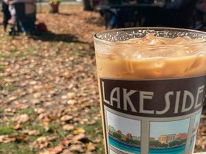 Iced coffee in a glass labeled "Lakeside" on an outdoor patio with autumn leaves and people in the background.