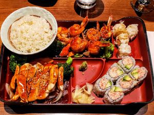 Bento box with rice, teriyaki chicken, shrimp, sushi rolls, shumai dumplings, broccoli, and ginger.