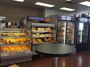 Pastry cases, chilled drinks, and sandwiches inside a simple, well-lit cafe with a round table in front.