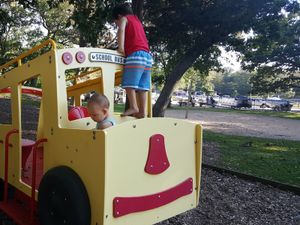 Two children play on a yellow school bus playground structure at Sandy Beach park, with boats in the background.