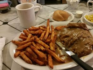 Plate of crispy fries, breaded meat with brown gravy, roll, corn, and coffee on a diner counter.