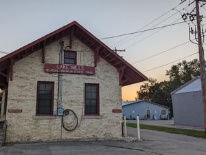 Old brick building with red trim labeled "Lake Mills Glacial Drumlin State Trail" at sunset.