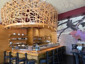 Sushi bar with wooden lattice ceiling feature, open shelving, and mural of a woman in a kimono on the wall.