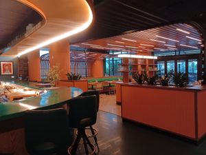 Modern restaurant interior with a curved green bar, orange paneling, plants, and sleek seating.
