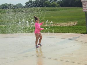 Young child in a bright pink swimsuit plays in arcing splash pad fountains on a sunny park day.