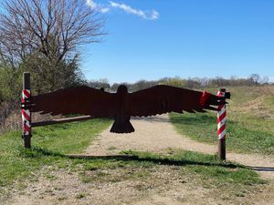 Metal gate shaped like a bird with outstretched wings blocks a gravel path at a grassy wildlife area.