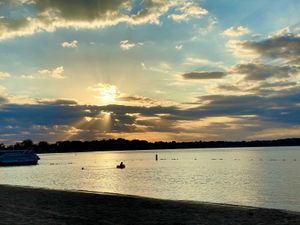 Sunbeams break through clouds over a calm lake, with a sandy shoreline and a lone boat and swimmer visible.