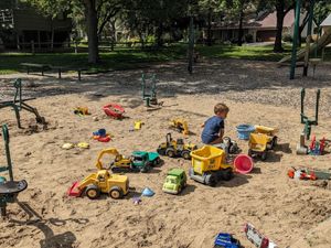 Young child playing with toy trucks and construction vehicles in a sandy playground area surrounded by trees.