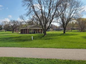 Open grassy park with leafless trees, a covered picnic shelter, and a paved walking path under a blue sky.