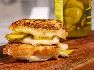 Toasted grilled cheese with melted white cheese and pickle slices on wooden board, jar of pickles in background