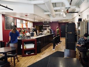 Tea shop interior with maroon service counter, illuminated menu boards, exposed white ductwork, wooden floors, seated…