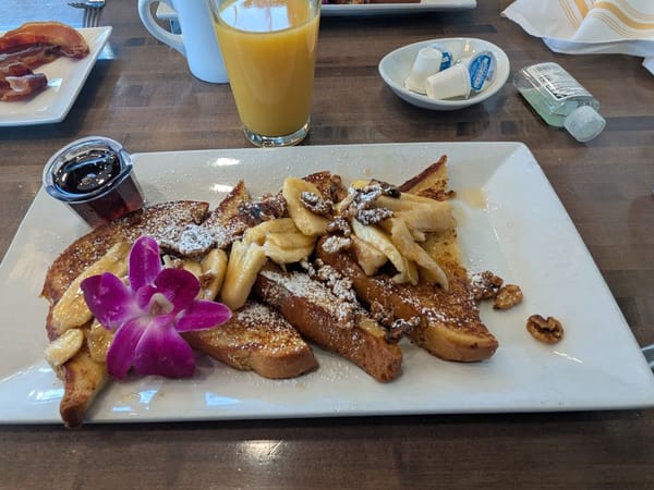 Golden pancakes topped with bananas, nuts, and a purple flower, served with syrup and orange juice.