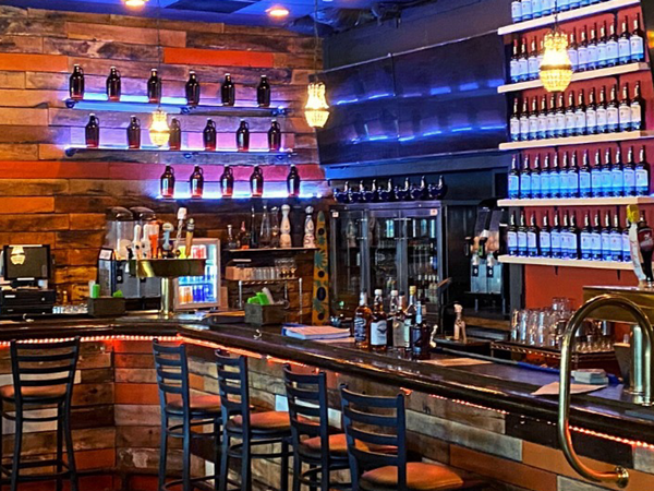 Rustic bar at Third Street Tavern with bottle-lined shelves, glowing lights, and wooden stools.