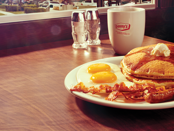 Plate of pancakes, eggs, bacon, and sausage with a Denny's mug on a sunny diner table.