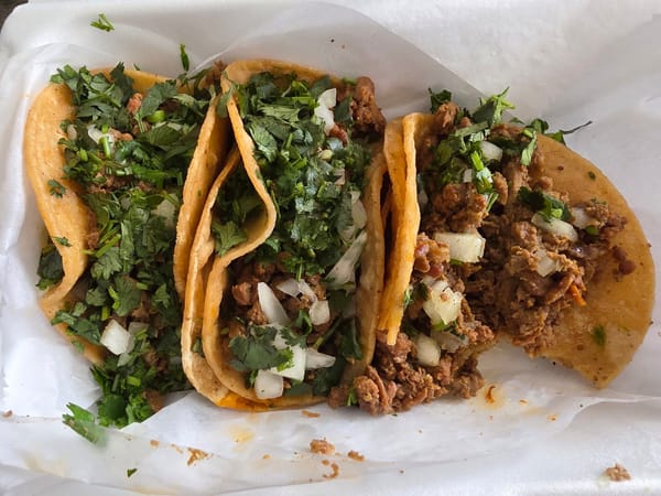 Three street-style beef tacos with onions and cilantro on corn tortillas, served on white paper.
