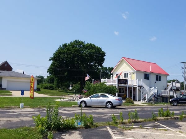 Bright yellow ice cream and grill shop with red roof, outdoor seating, parking, and an "open" flag.