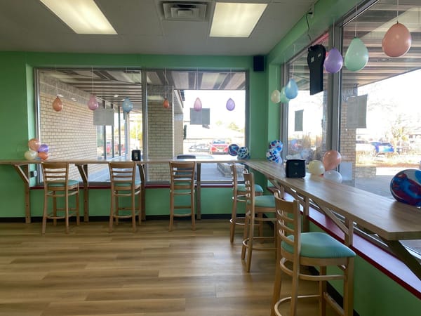 Sunlit ice cream shop with pastel balloons, wooden chairs, and a long counter overlooking large windows.