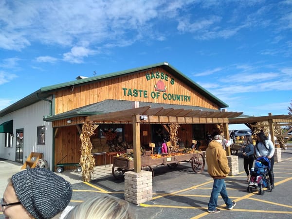 People visit a rustic farm market decorated with autumn produce and cornstalks under a bright blue sky.