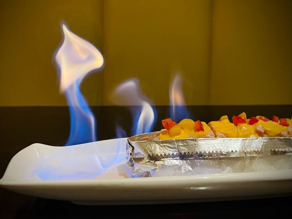 Sushi rolls topped with sauce and diced peppers served flaming on a white plate at Kanpai Izakaya.