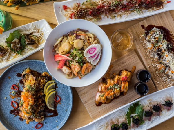 Colorful ramen bowl, assorted sushi rolls, and sashimi plates arranged on a wooden table with sauces.