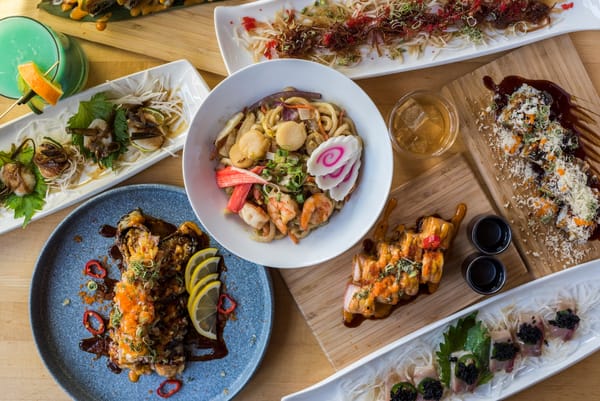 Colorful Japanese dishes, including noodles, skewers, and sushi, arranged on a wooden table with drinks.