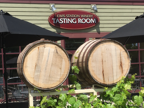 Two large wooden barrels and green vines outside Lewis Station Winery Tasting Room with umbrellas and seating.