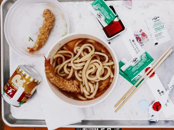 Bowl of udon noodle soup with tempura shrimp, chopsticks, soy sauce packets, and a can of Japanese drink on a tray.
