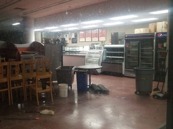 Dimly lit cafe interior with stacked chairs, cleaning supplies, and empty display cases near a counter.