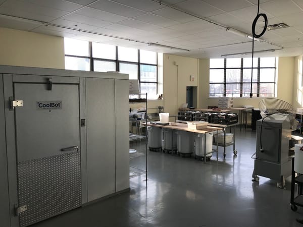 Spacious bakery kitchen with large windows, stainless steel equipment, and a walk-in cooler at ORIGIN Breads.