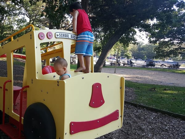 Two children play on a yellow school bus playground structure at Sandy Beach park, with boats in the background.