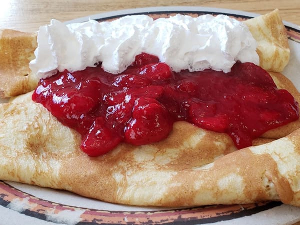 Golden crepes topped with strawberry sauce and a generous swirl of whipped cream on a diner plate.