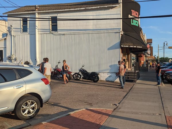 People stand outside El Taco Loco restaurant on a sunny day, with motorcycles and cars parked nearby.