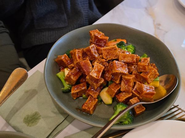 Glazed tofu cubes served over steamed broccoli and vegetables in a gray bowl, with a serving spoon.